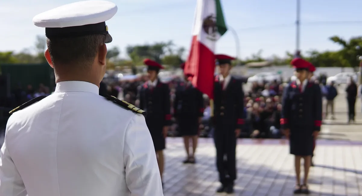 Back view of a white-dress military officer addressing a line of cadets at a ceremony, with a Mexican flag in the background.