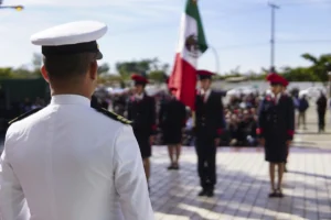 Back view of a white-dress military officer addressing a line of cadets at a ceremony, with a Mexican flag in the background.