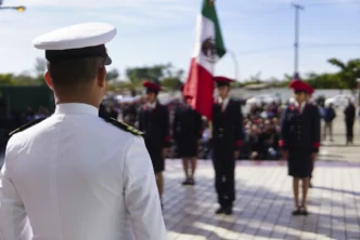 Back view of a white-dress military officer addressing a line of cadets at a ceremony, with a Mexican flag in the background.