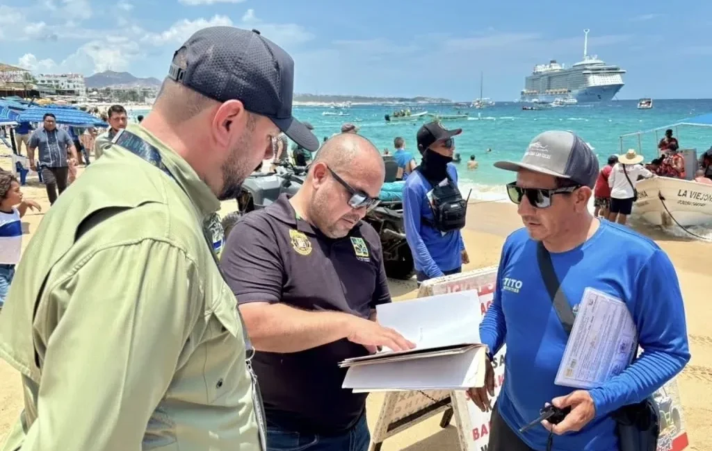Three men on a sunny beach examine documents, with a cruise ship and turquoise water in the background.