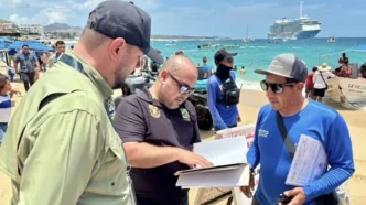 Three men on a sunny beach examine documents, with a cruise ship and turquoise water in the background.