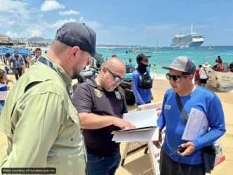 Three men on a sunny beach examine documents, with a cruise ship and turquoise water in the background.
