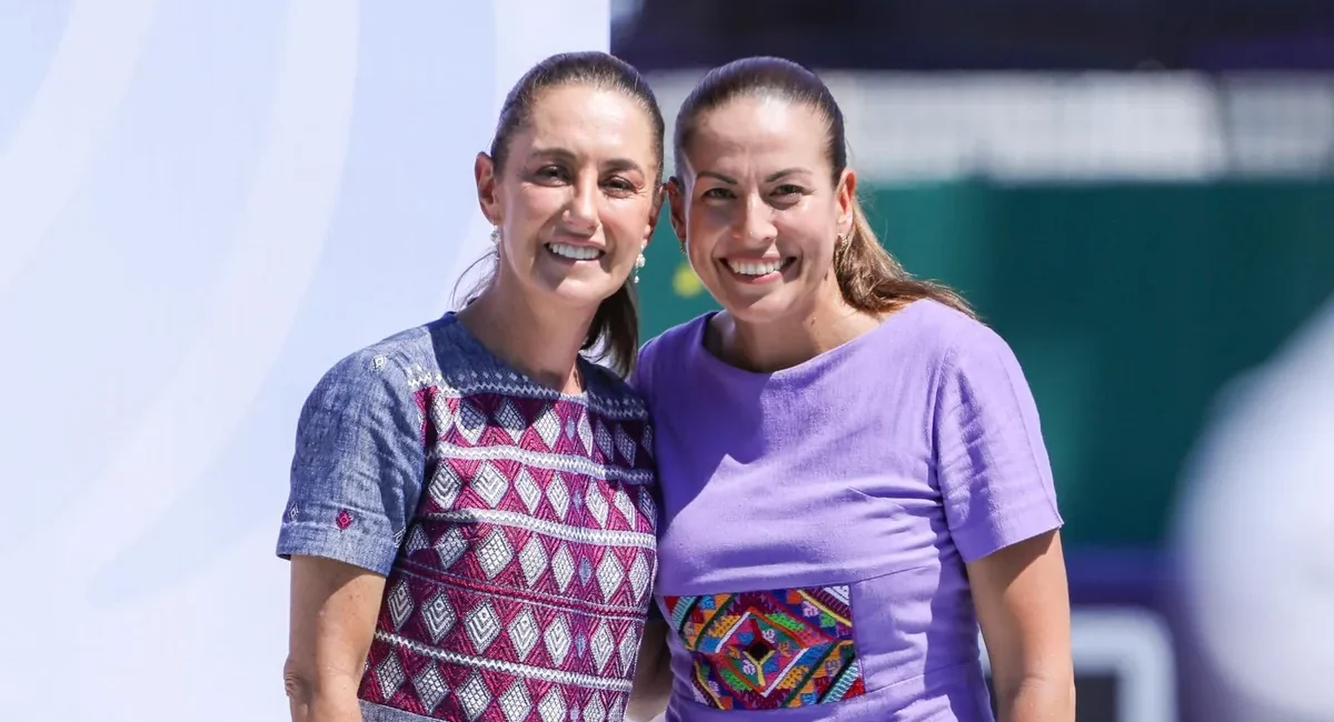 Two smiling women stand close together, one in a patterned blue dress and the other in a lavender shirt with a colorful embroidery patch, posing outdoors.