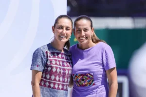 Two smiling women stand close together, one in a patterned blue dress and the other in a lavender shirt with a colorful embroidery patch, posing outdoors.
