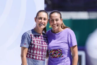 Two smiling women stand close together, one in a patterned blue dress and the other in a lavender shirt with a colorful embroidery patch, posing outdoors.