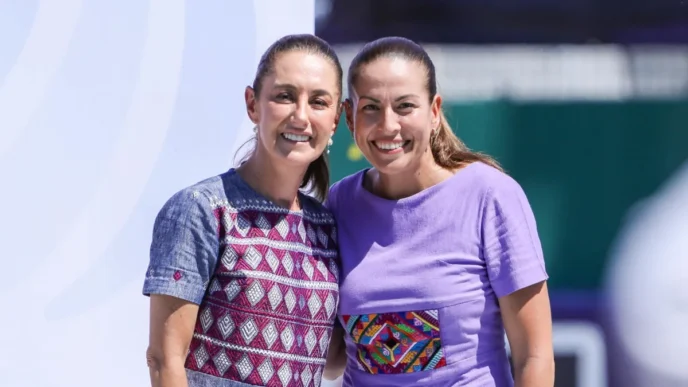 Two smiling women stand close together, one in a patterned blue dress and the other in a lavender shirt with a colorful embroidery patch, posing outdoors.