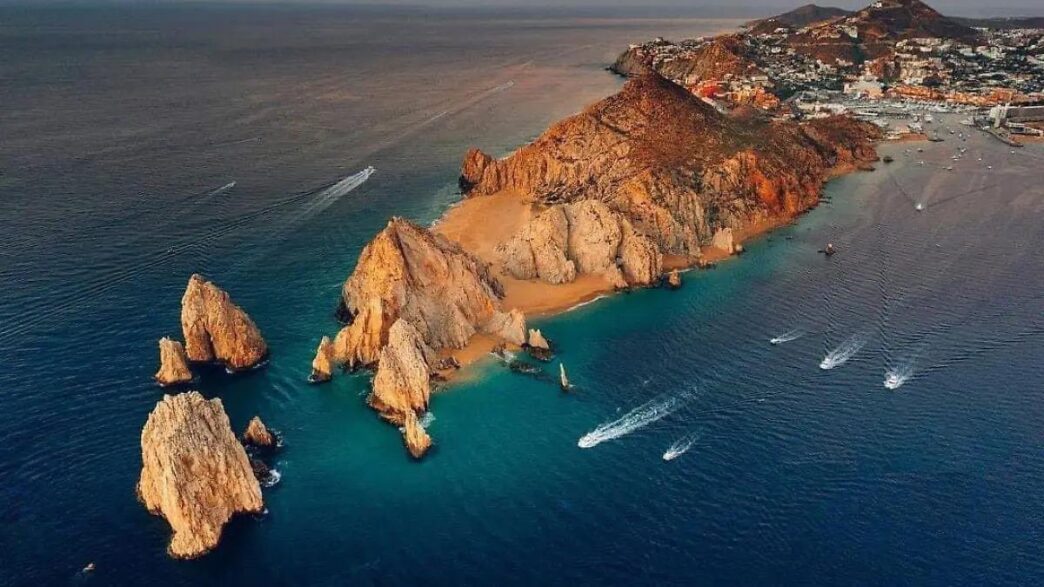 CaboSanLucas-1 - Gringo Gazette A rocky coastal headland with orange cliffs extending into blue-green water; several boats leave white wake trails nearby in a calm sea.