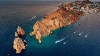 CaboSanLucas-1 - Gringo Gazette A rocky coastal headland with orange cliffs extending into blue-green water; several boats leave white wake trails nearby in a calm sea.