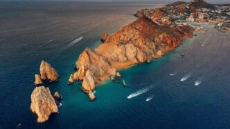 A rocky coastal headland with orange cliffs extending into blue-green water; several boats leave white wake trails nearby in a calm sea.
