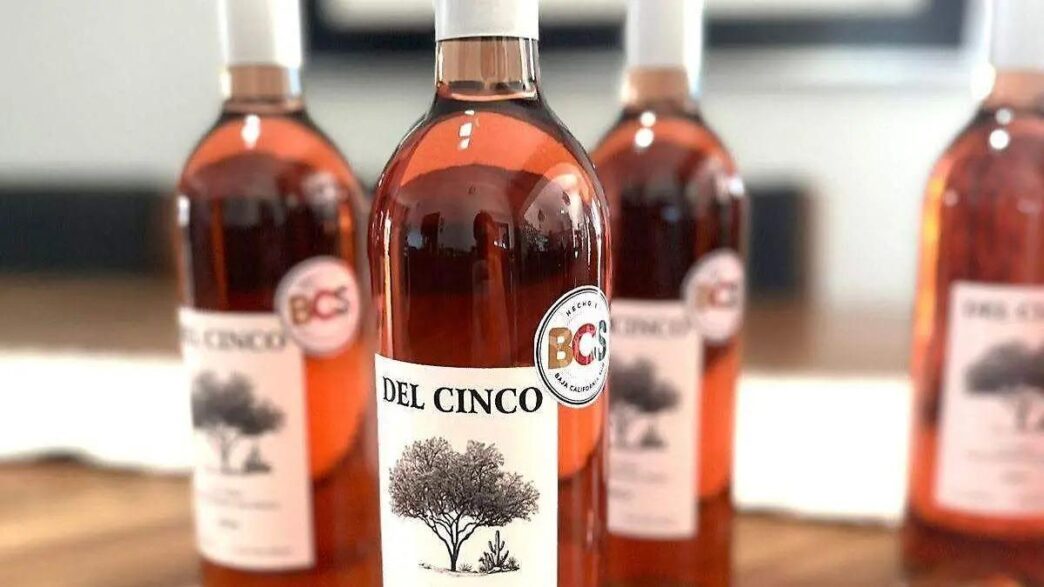 Comandu - Gringo Gazette Del Cinco bottle in focus with a tree label, three blurred bottles behind on a wooden surface.