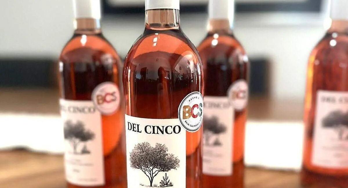 Del Cinco bottle in focus with a tree label, three blurred bottles behind on a wooden surface.