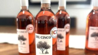 Del Cinco bottle in focus with a tree label, three blurred bottles behind on a wooden surface.
