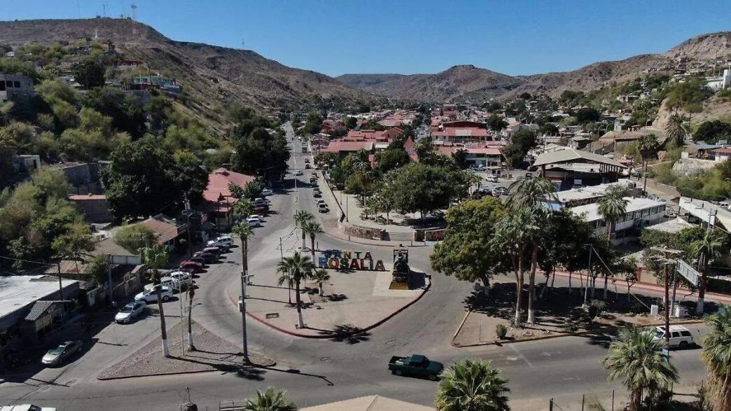 Earthquake - Gringo Gazette Aerial view of a desert town center with a roundabout, palm trees, and a large 'ROSALIA' sign in the plaza, surrounded by low hills and red-roofed buildings.