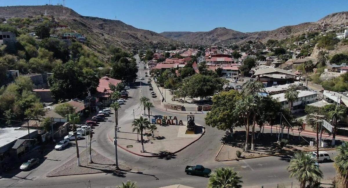 Aerial view of a desert town center with a roundabout, palm trees, and a large 'ROSALIA' sign in the plaza, surrounded by low hills and red-roofed buildings.