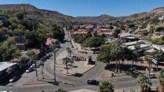 Earthquake - Gringo Gazette Aerial view of a desert town center with a roundabout, palm trees, and a large 'ROSALIA' sign in the plaza, surrounded by low hills and red-roofed buildings.