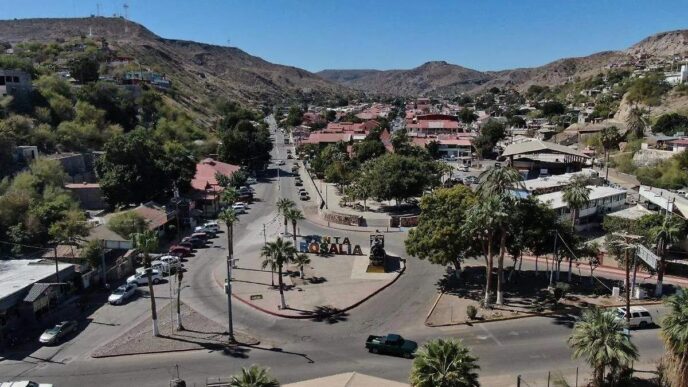 Aerial view of a desert town center with a roundabout, palm trees, and a large 'ROSALIA' sign in the plaza, surrounded by low hills and red-roofed buildings.