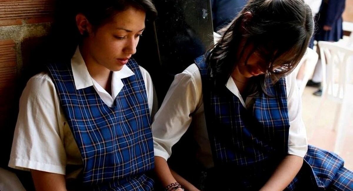 Two girls in blue plaid school uniforms sit side by side against a brick wall, looking at a tablet together.