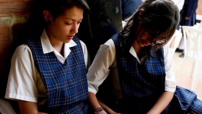 Two girls in blue plaid school uniforms sit side by side against a brick wall, looking at a tablet together.