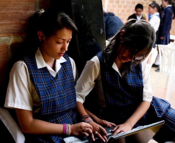 Education - Gringo Gazette Two girls in blue plaid school uniforms sit side by side against a brick wall, looking at a tablet together.