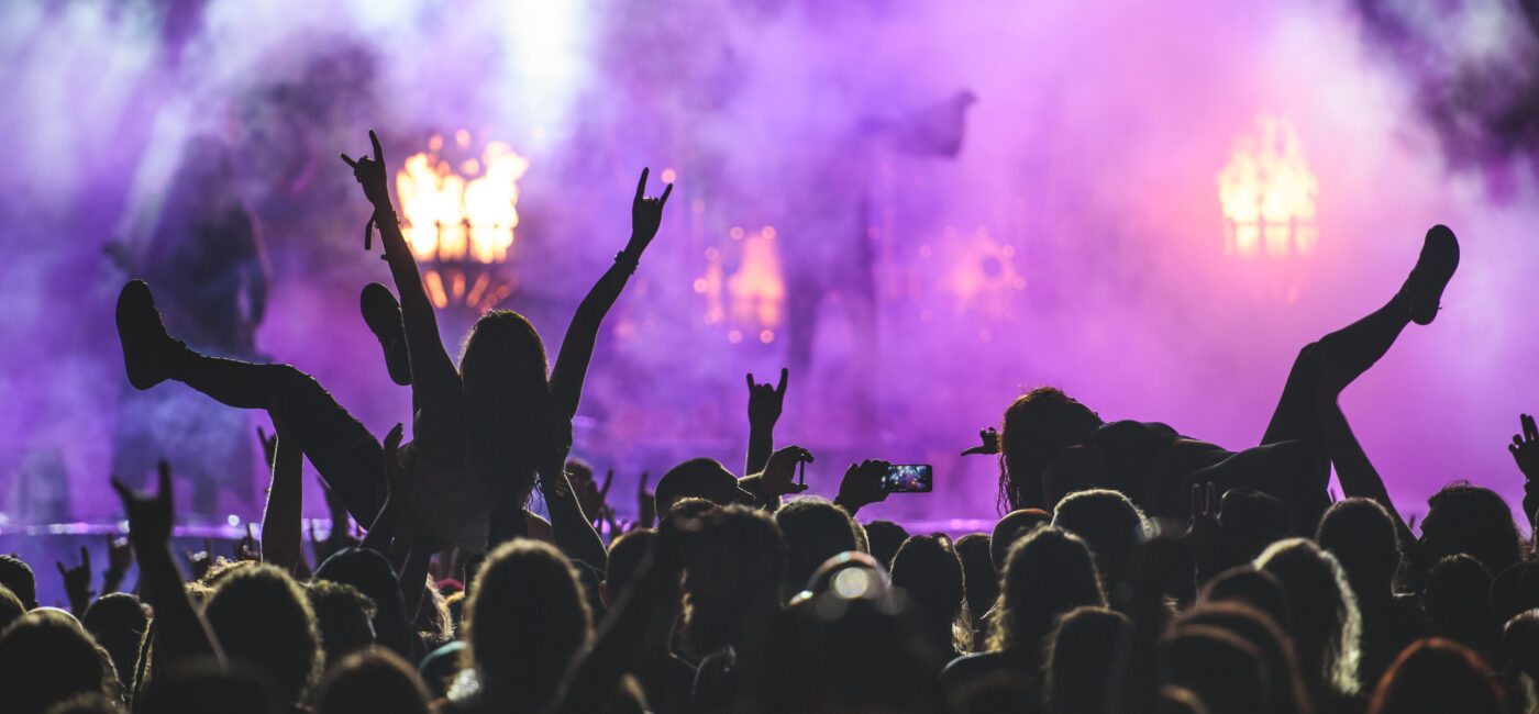 Concert crowd with raised hands silhouetted against purple stage lights and smoke.