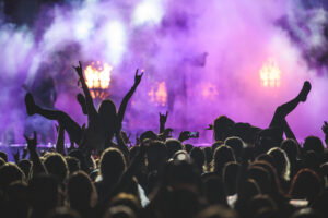 Concert crowd with raised hands silhouetted against purple stage lights and smoke.