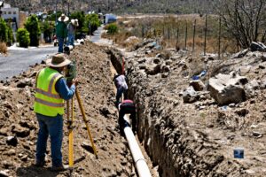 Construction crew trenching a long ditch to lay a large pipe, with a surveyor using a level nearby.