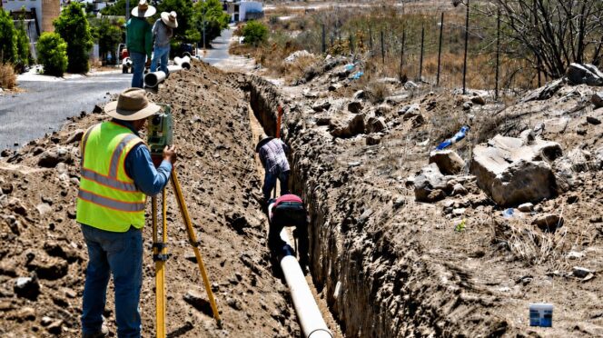 Construction crew trenching a long ditch to lay a large pipe, with a surveyor using a level nearby.