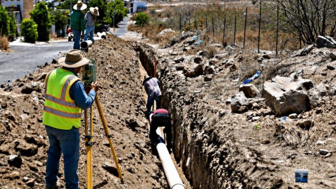 Construction crew trenching a long ditch to lay a large pipe, with a surveyor using a level nearby.