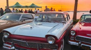Vintage cars line a waterfront lot at sunset, with a light blue and red two-tone classic in the foreground and a crowd behind them.