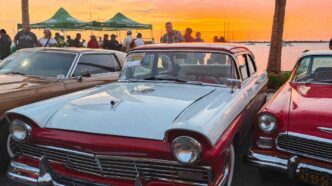 LaPazCarShow01 - Gringo Gazette Vintage cars line a waterfront lot at sunset, with a light blue and red two-tone classic in the foreground and a crowd behind them.