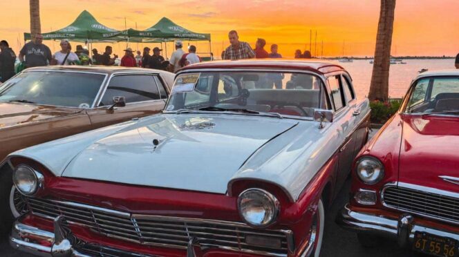 Vintage cars line a waterfront lot at sunset, with a light blue and red two-tone classic in the foreground and a crowd behind them.