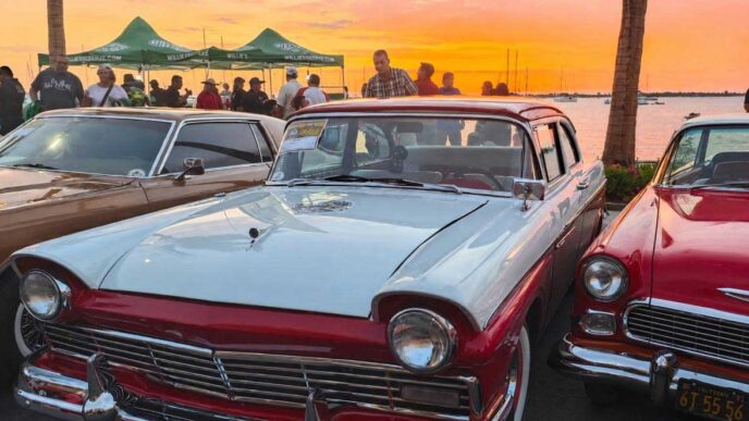 Vintage cars line a waterfront lot at sunset, with a light blue and red two-tone classic in the foreground and a crowd behind them.