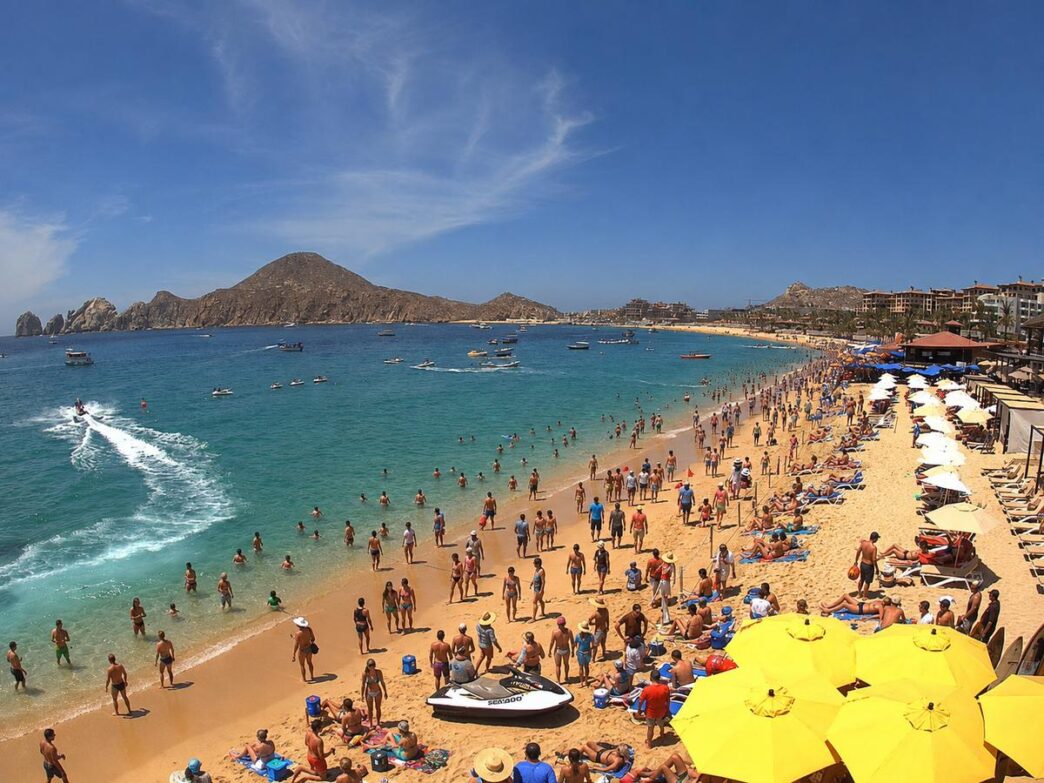 Los-Cabos-1 - Gringo Gazette Crowded golden-sand beach with bright yellow umbrellas, sunbathers, and swimmers under a blue sky.