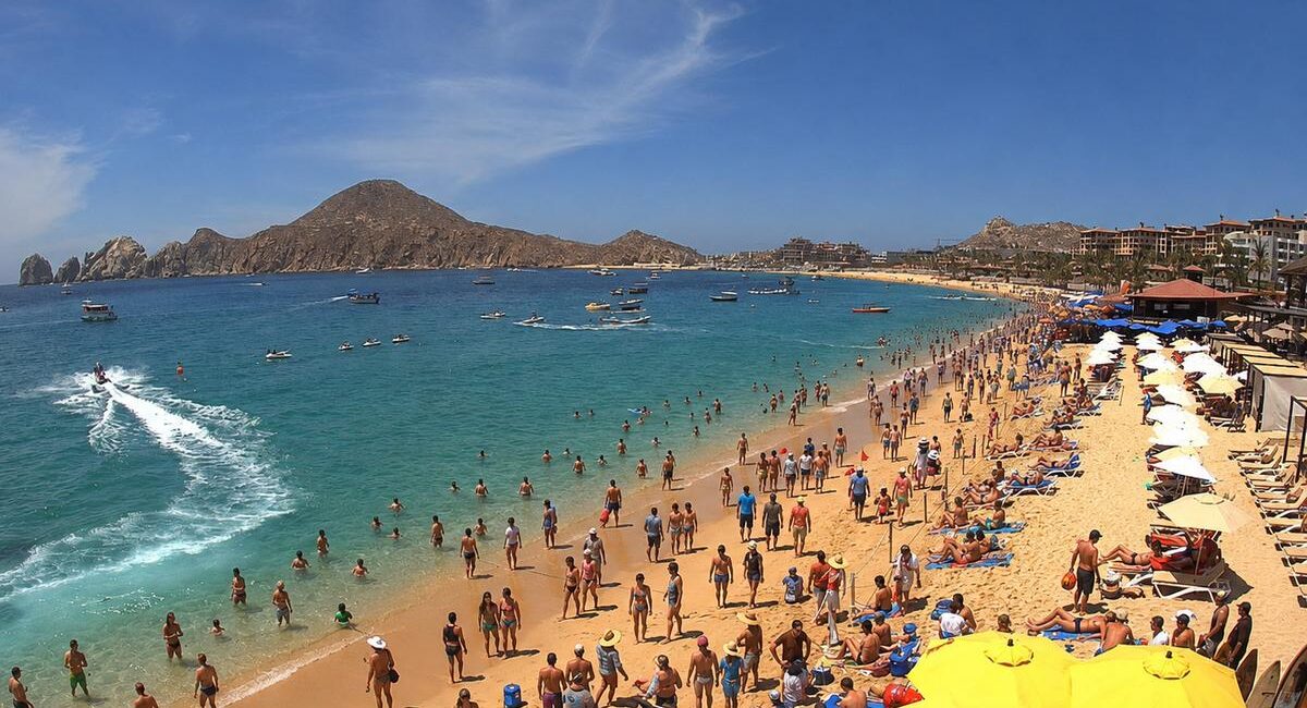 Crowded golden-sand beach with bright yellow umbrellas, sunbathers, and swimmers under a blue sky.