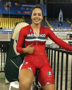 Female Mexican cyclist in a red Mexico kit gives a thumbs up inside a stadium, seated on a stationary bike with a Mexican flag patch and Olympic rings on her shorts.