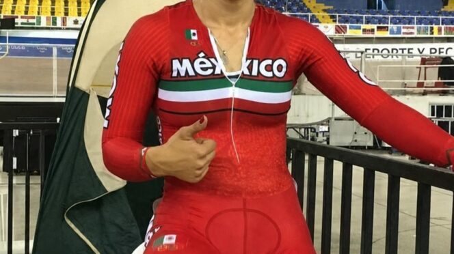 Female Mexican cyclist in a red Mexico kit gives a thumbs up inside a stadium, seated on a stationary bike with a Mexican flag patch and Olympic rings on her shorts.