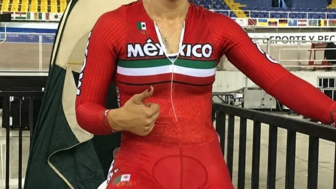 Female Mexican cyclist in a red Mexico kit gives a thumbs up inside a stadium, seated on a stationary bike with a Mexican flag patch and Olympic rings on her shorts.