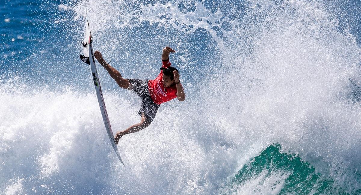 Surfer in a red shirt launches off a breaking wave, spraying water in all directions as a teal-green wave roars nearby.