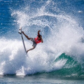 Surfer in a red shirt launches off a breaking wave, spraying water in all directions as a teal-green wave roars nearby.