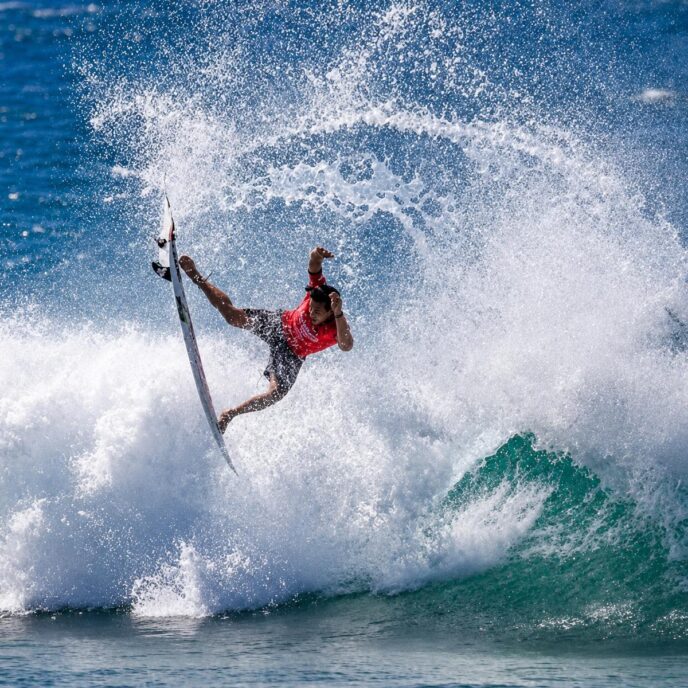 Surfer in a red shirt launches off a breaking wave, spraying water in all directions as a teal-green wave roars nearby.