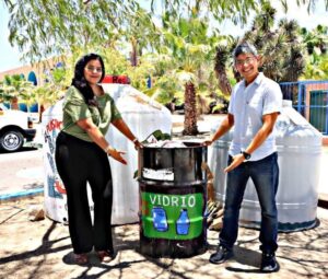 Two people stand beside a glass recycling bin labeled 'VIDRIO' showing bottles, outdoors with palm trees in the background.