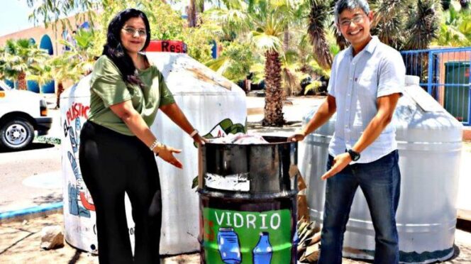 Two people stand beside a glass recycling bin labeled 'VIDRIO' showing bottles, outdoors with palm trees in the background.