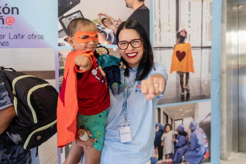 Smiling woman in a light blue shirt holds a young boy in an orange cape and mask, both pointing toward the camera. Behind them are event banners.