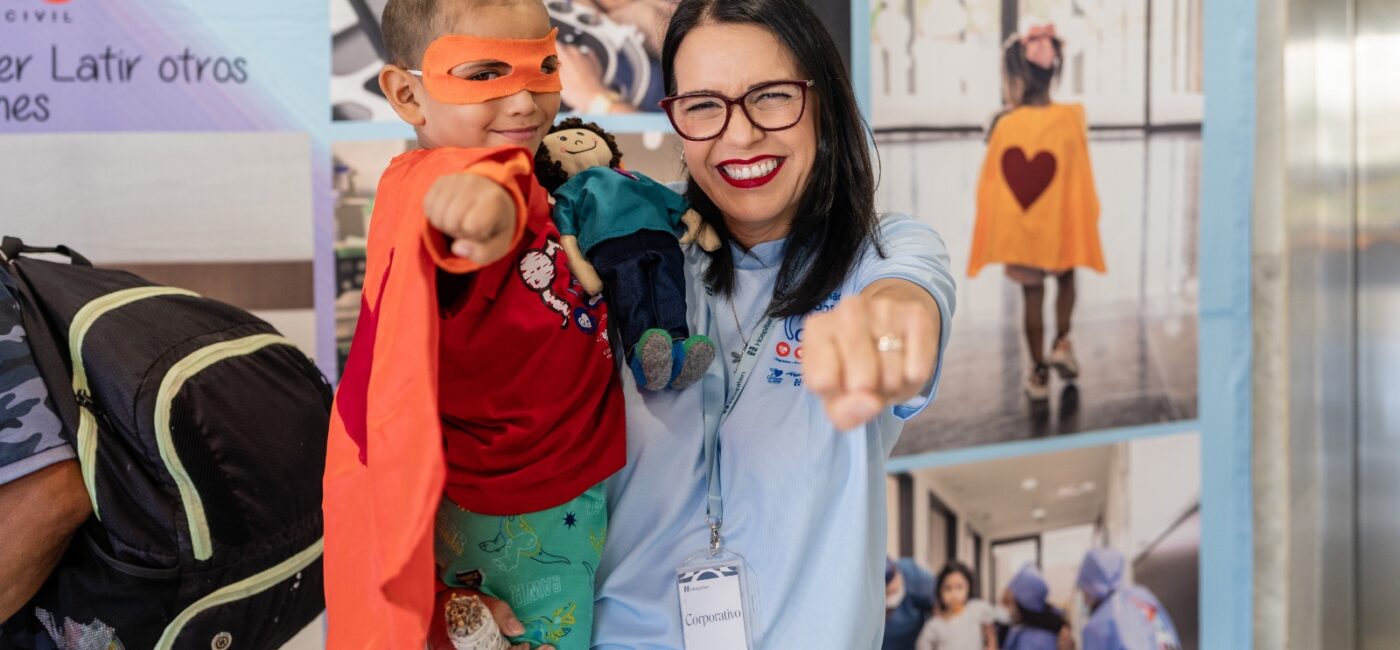 Smiling woman in a light blue shirt holds a young boy in an orange cape and mask, both pointing toward the camera. Behind them are event banners.