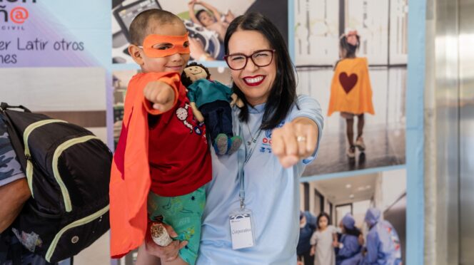 Smiling woman in a light blue shirt holds a young boy in an orange cape and mask, both pointing toward the camera. Behind them are event banners.