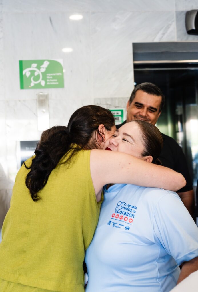 Two women embrace warmly in a public space, one in a yellow top and the other in a light blue shirt with a 'Unidos de Corazón' logo, a man smiles in the background.