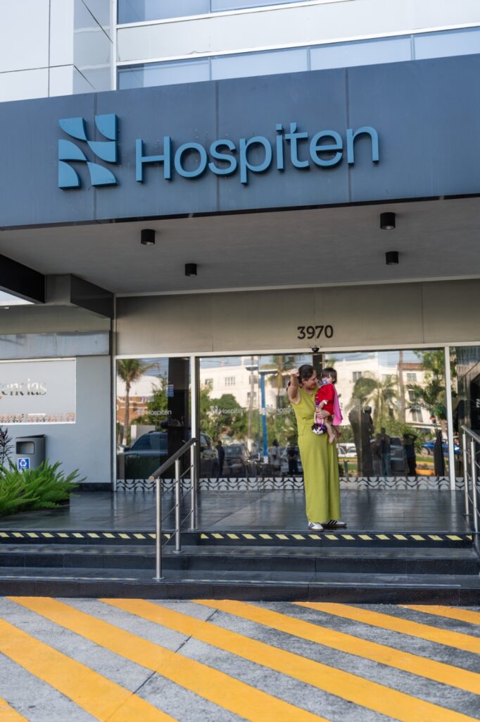 Woman in a lime dress holding a child outside a hospital entrance with a blue 'Hospiten' sign above.