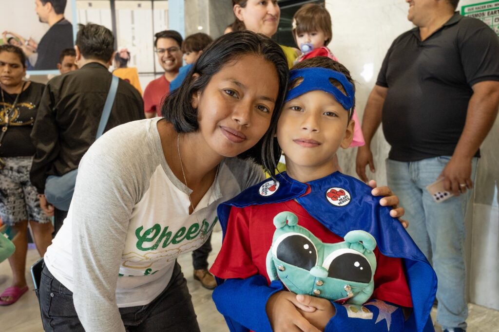 Woman with short dark hair leaning in and smiling beside a boy in a blue superhero cape and mask at a crowded indoor event.