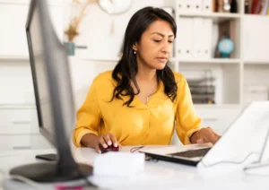 Woman in a yellow blouse sits at a white desk, using a computer mouse while working on a tablet in an office setting.