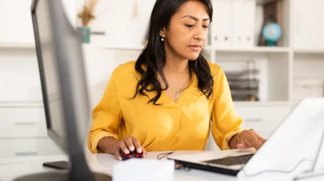 Woman in a yellow blouse sits at a white desk, using a computer mouse while working on a tablet in an office setting.